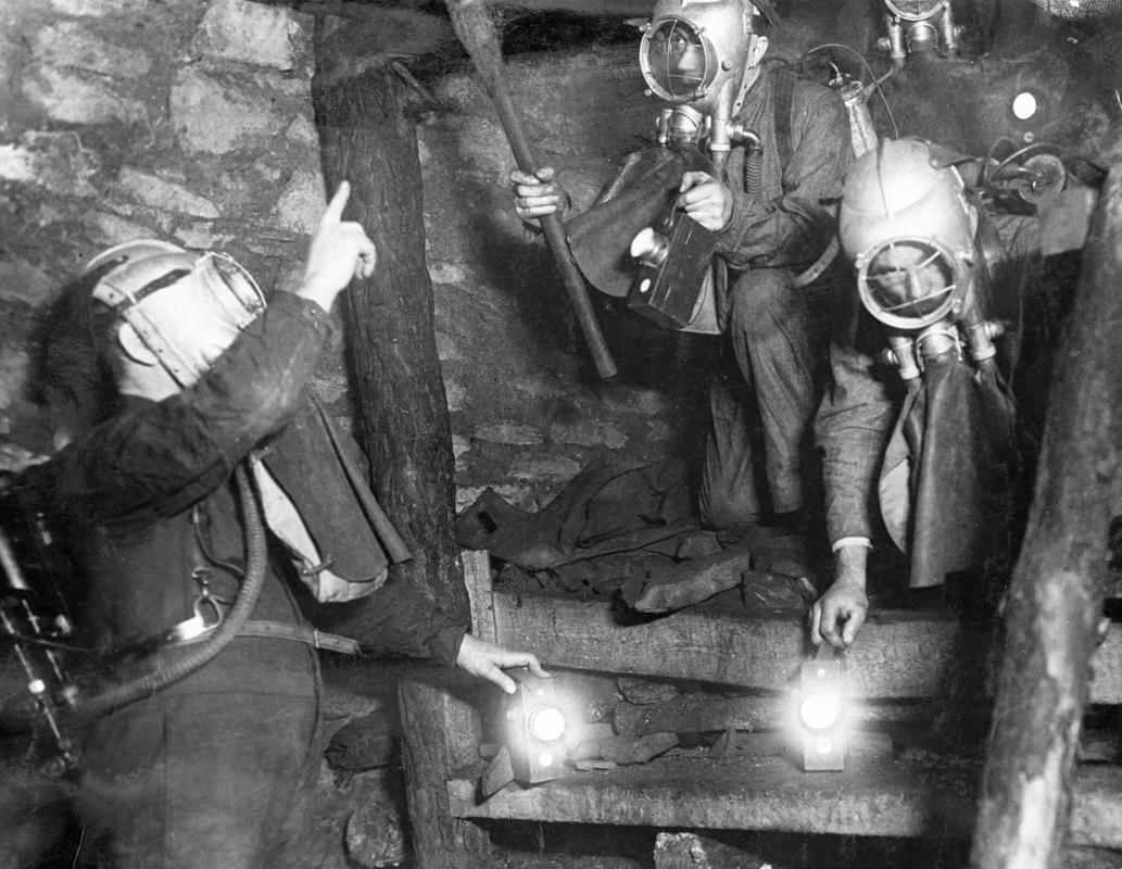 Brigade men, from Dinas Rescue Station, in Draegar breathing apparatus, practising in model underground gallery, simulating an exploring party climbing over fallen roofing. They are carrying electric inspection lamps. Instructor on left. Man second from left is testing the roof for danger spots with a mandrel helve.