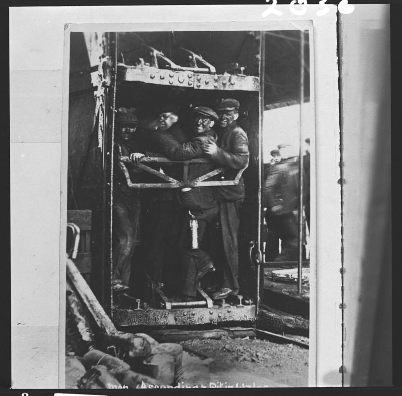 Black and white film negative of a photograph showing four men in a cage after arriving back on the surface, Pandy Pit, Penygraig.