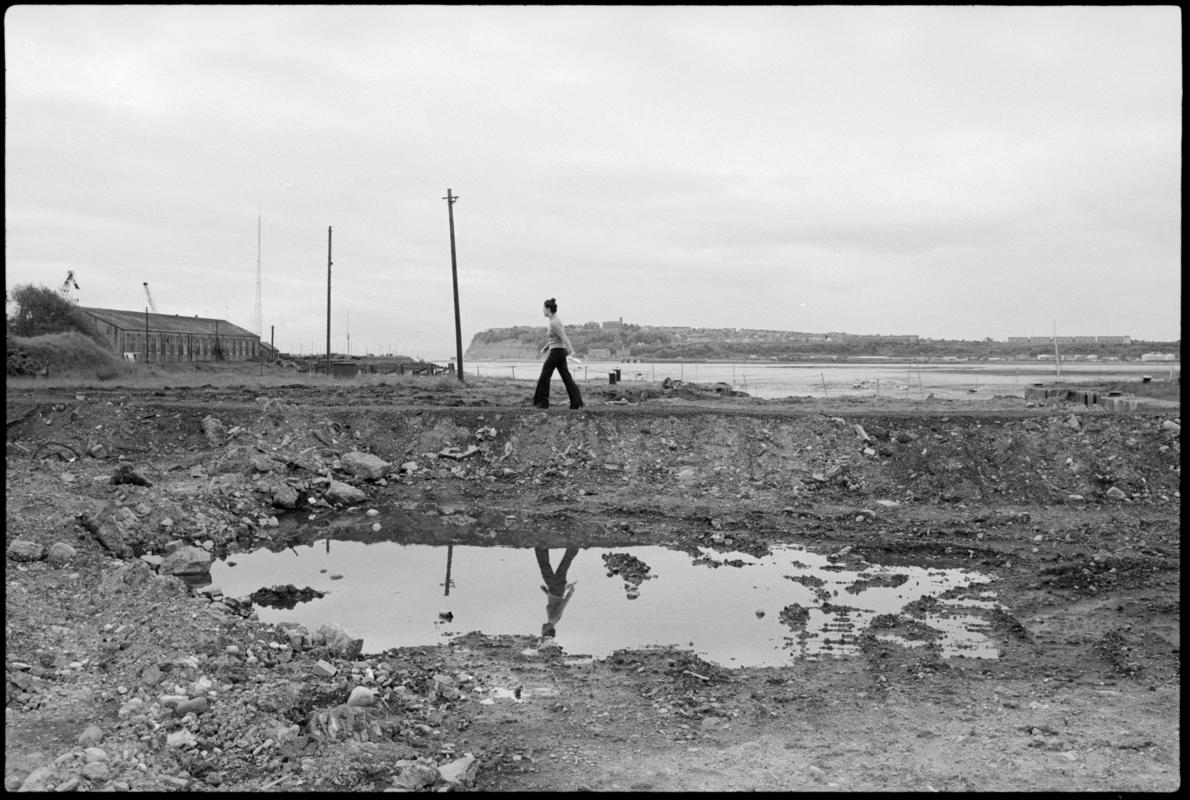 A walking figure reflected in a pool in the filled in Bute East Dock basin. Penarth head in the background.