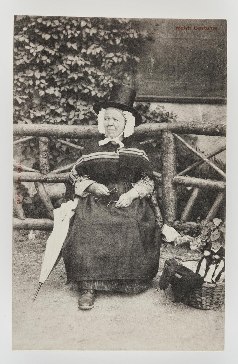 Woman in Welsh costume knitting in garden - umbrella on her arm; wool &amp; Welsh dolls in basket beside her.