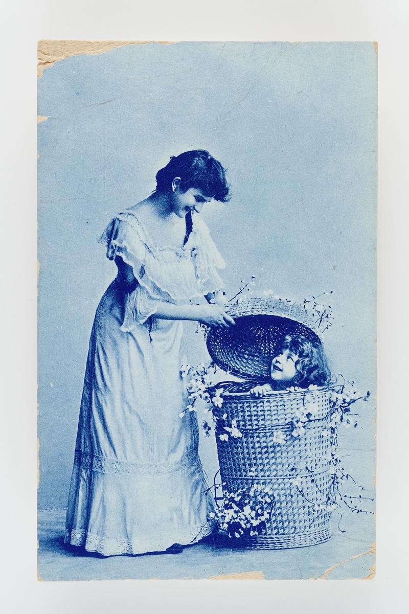 Photograph of a woman in Edwardian dress with child in basket.  Postmark:  Newquay, Cards.