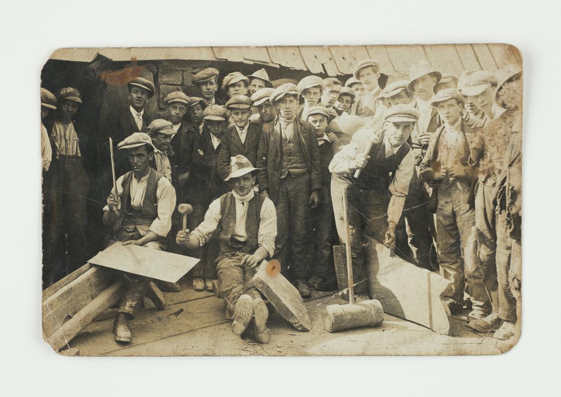Three quarrymen demonstrating the skill of slate splitting and dressing against a background of about 27 quarrymen. Said to have been taken somewhere at Dinorwig Quarry during the 1920s.