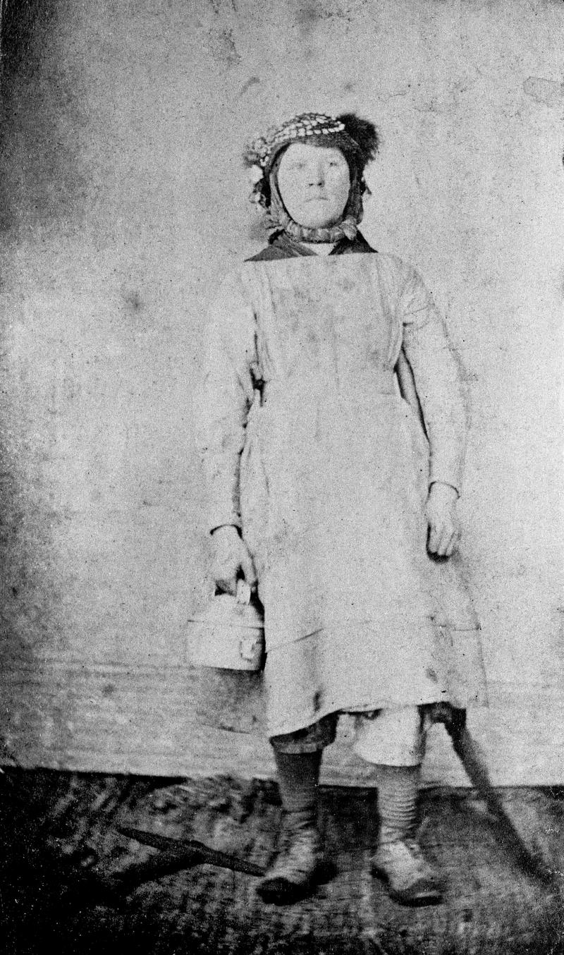 Girl emplyed on surface at a colliery or at an ironworks, holding a food tin in right hand with a crossed mandrel and hammer on floor to her right.