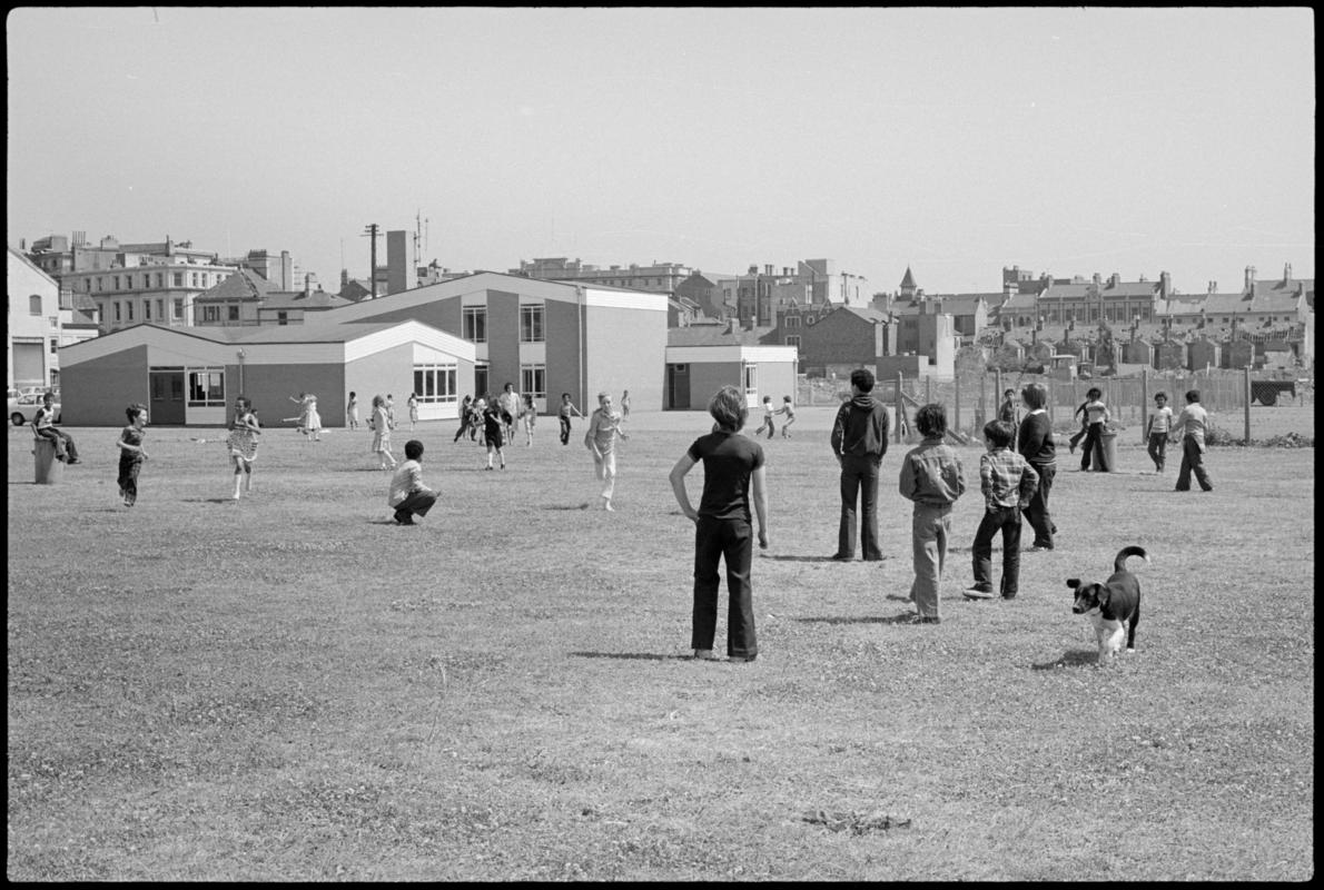Children playing in the large grass playground surrounding Mountstuart Primary School, Adelaide Street, Butetown. School visible in the background.
