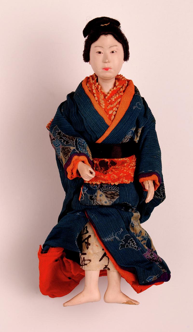 &#039;Sakura&#039; type Japanese doll (standing); head, hands and lower legs made of wood covered with gofum of a fleshy colour; intricate coiffure tied with ribbons; narrow black eyes; right hand is held forward, and probably held a parasol; costume is a patterne