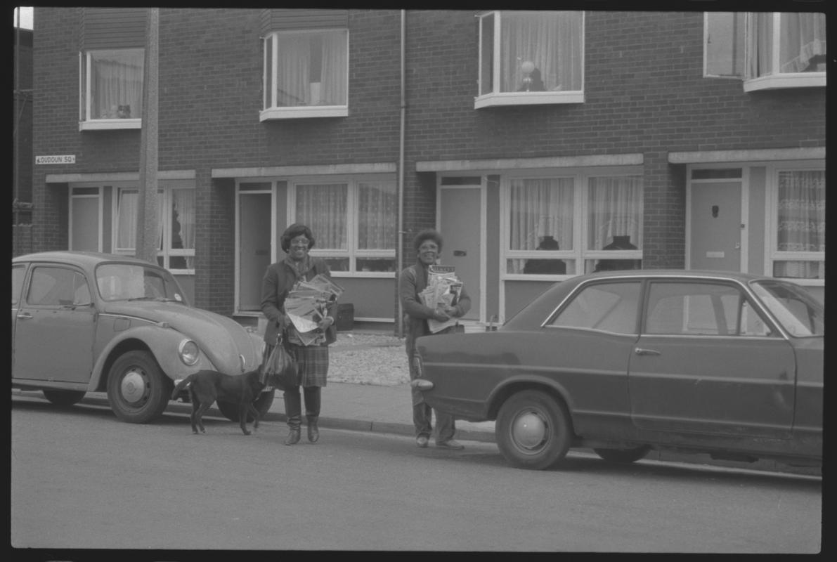 Cars parked outside modern terraced housing at Loudoun Square, Butetown. Two women carrying parcels are walking between them.