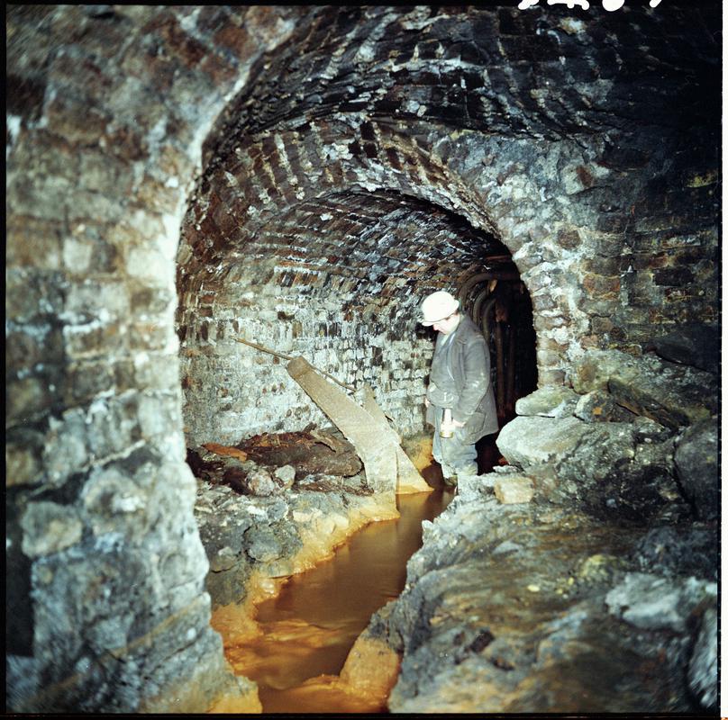 Colour film negative showing a mine official examining a section of a water balance machine cage, near the bottom of the Forge Pit, Blaenavon, 1975.  A slip of paper is attached to one edge of the negative.