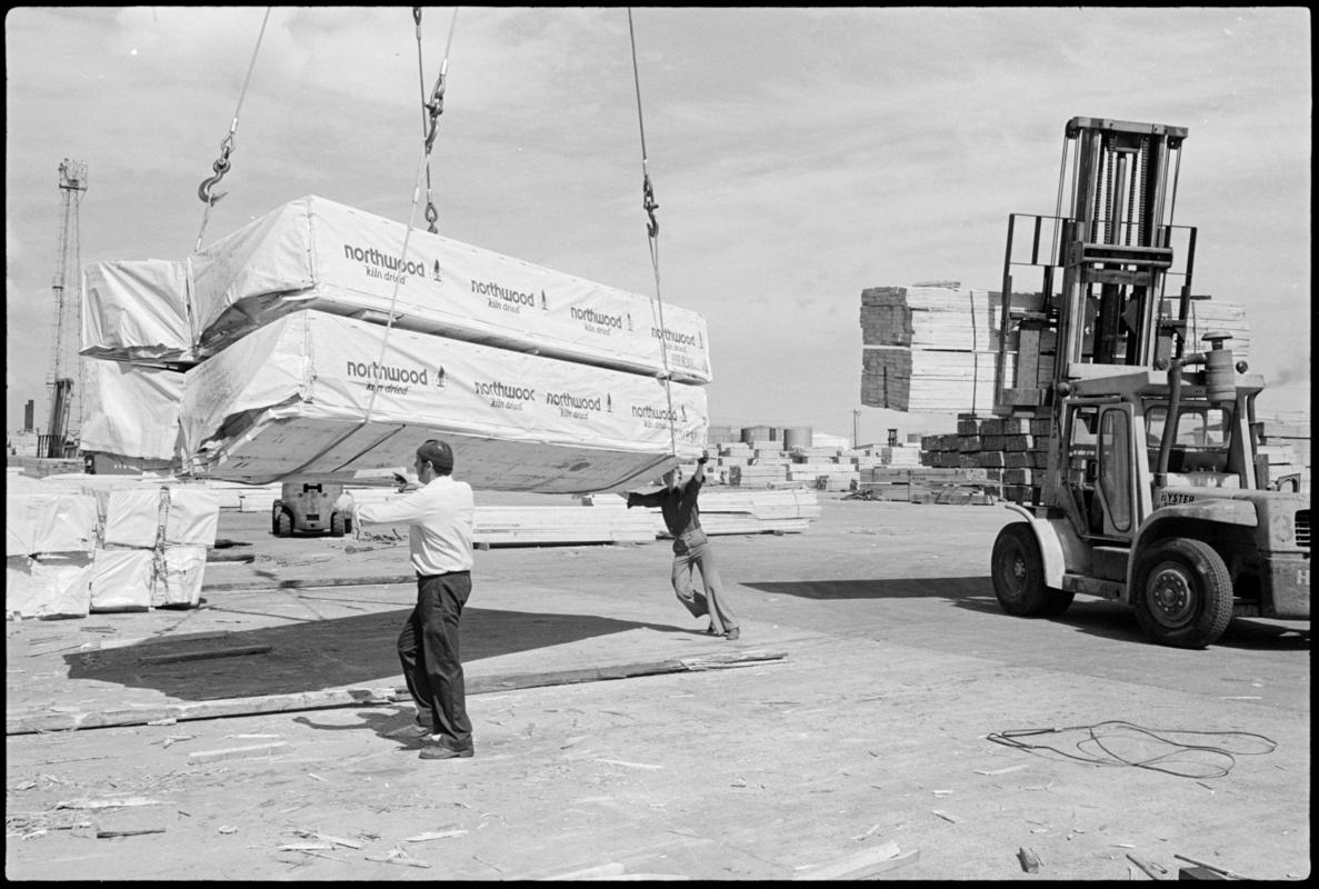 Two dockers steadying a crane load of timber to the ground, as large fork lift truck takes away a similar load for storage, Cardiff Docks.