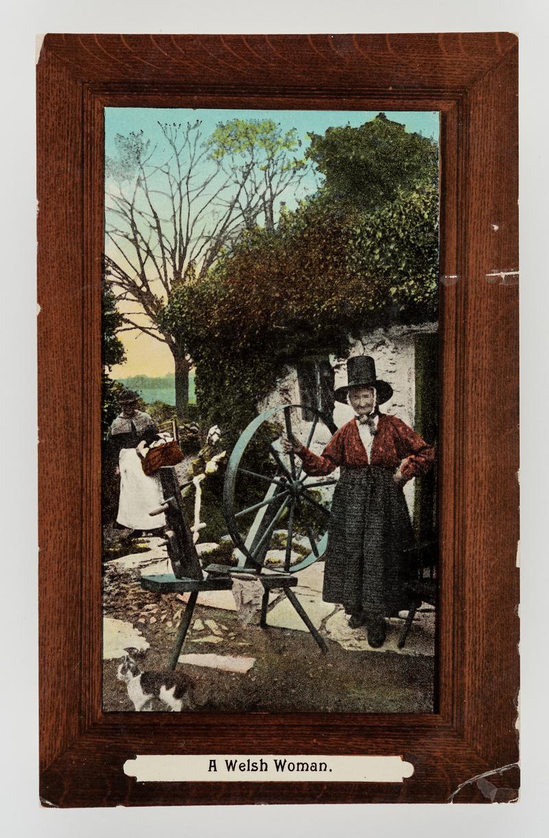 A Welsh woman (Ellen Lloyd, Bettws-y-Coed), in costume standing by spinning wheel outside cottage, with another lady approaching.
