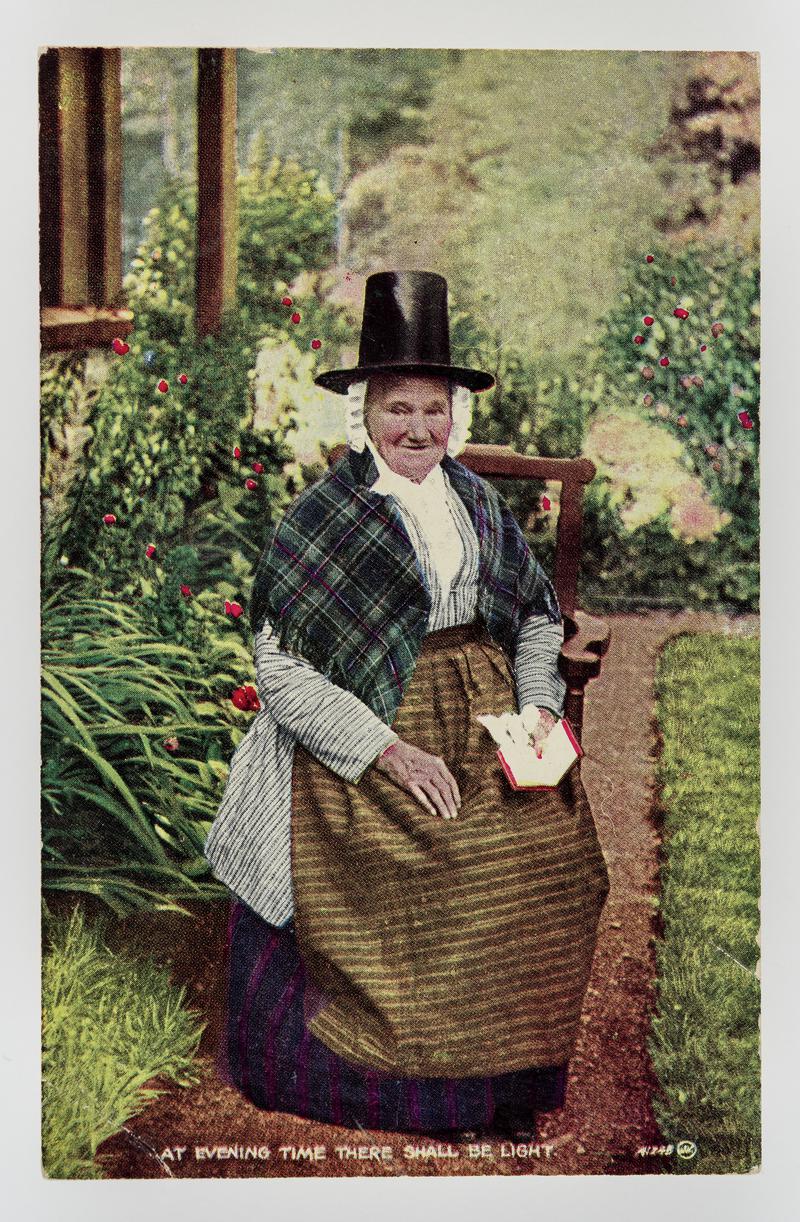Elderly lady in Welsh costume sitting in garden with book. Inscribed &#039;At evening time there shall be light.&#039;