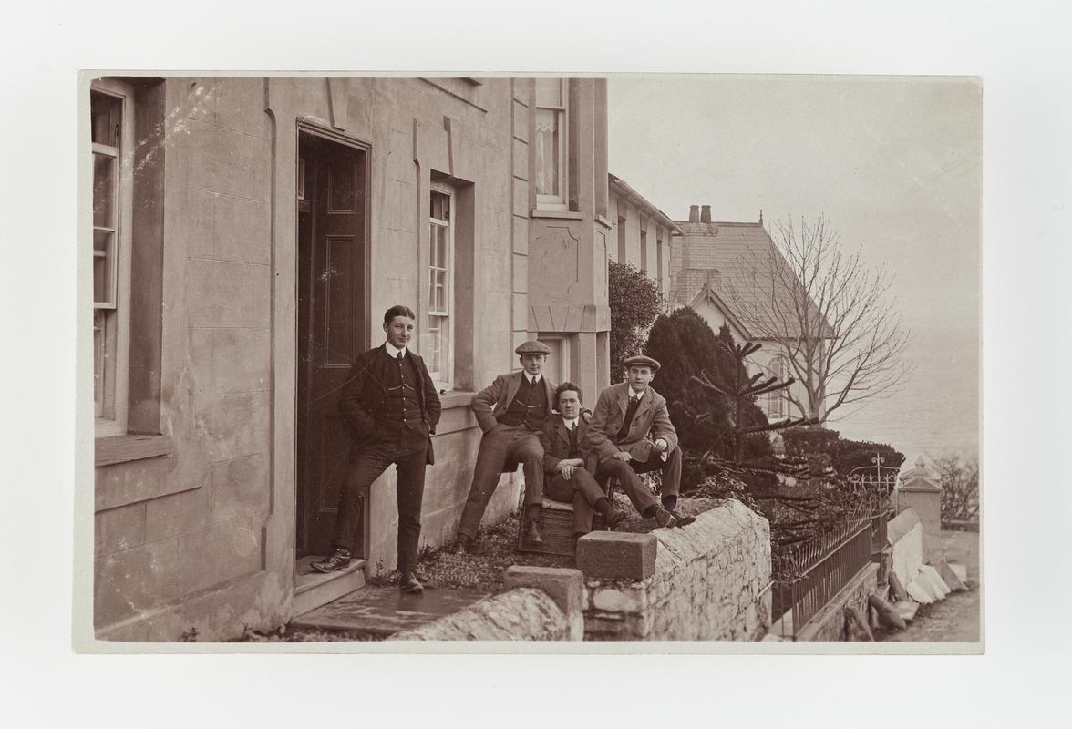 Photograph of four men outside a house.  All wearing suits, two also wearing flat caps.