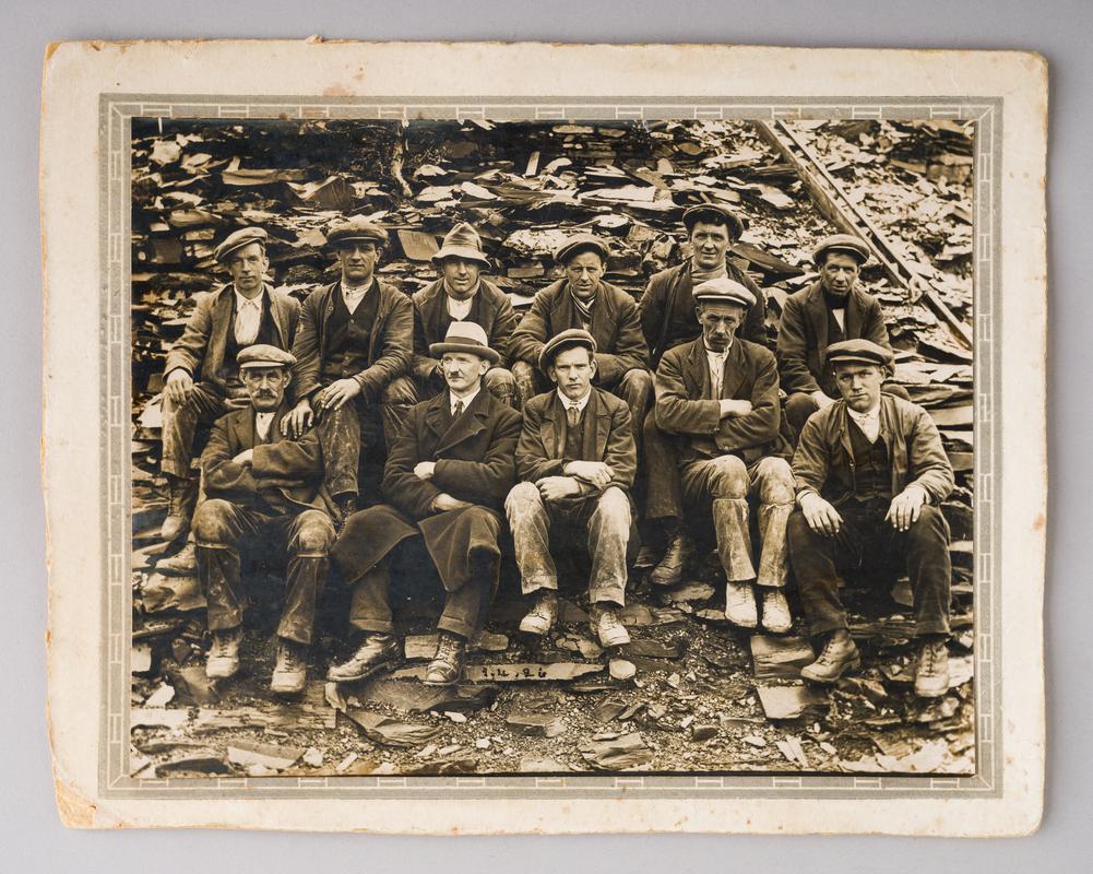 Group photograph of 11 Penrhyn quarry workers. Date &#039;1.4.26&#039; painted on slate in front of workers.
