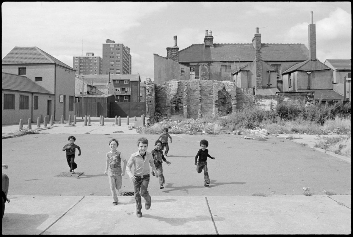 Children in Butetown running towards camera. New and derelict housing, including Loudoun Square flats, in the background.