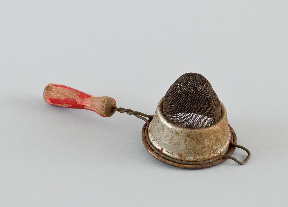 Metal tea strainer, with wooden handle attached by wire, red paint flaking off handle. Severe areas of rust visible.