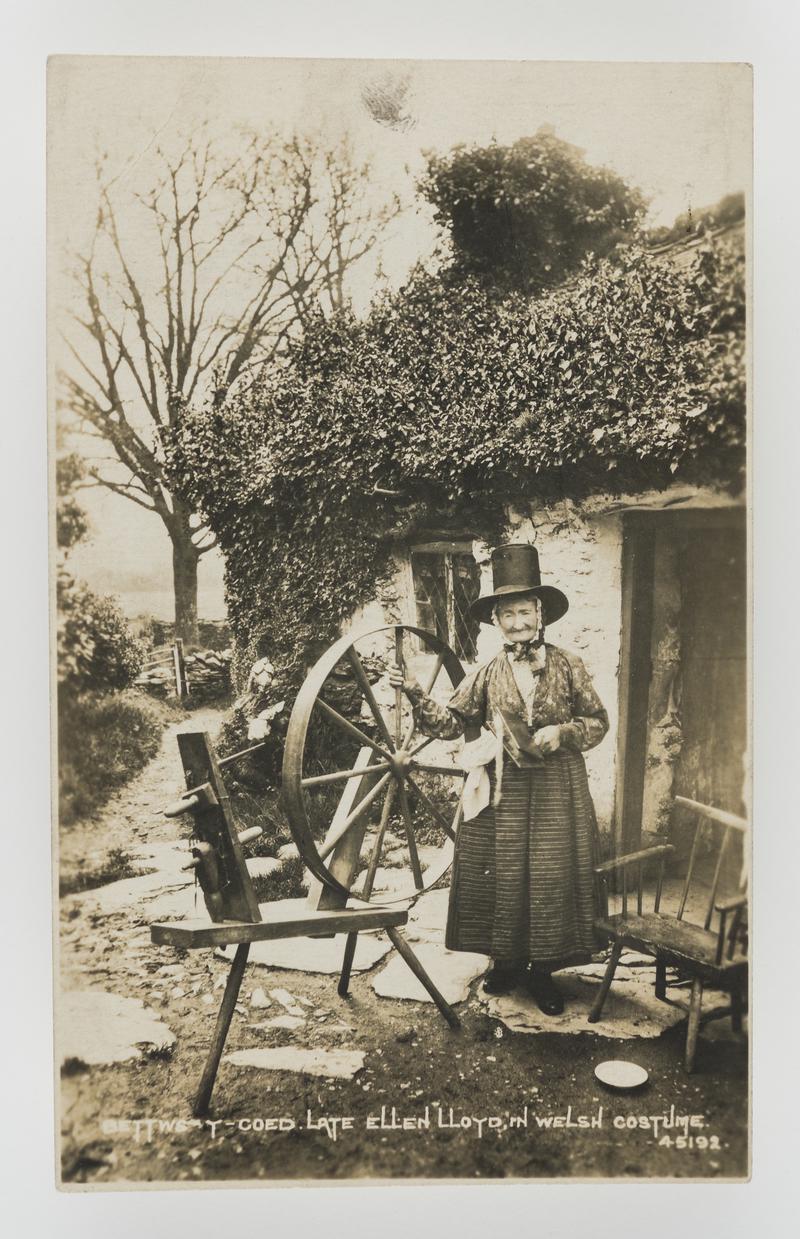 Ellen Lloyd, Bettws-y-Coed, in Welsh costume standing by spinning wheel.