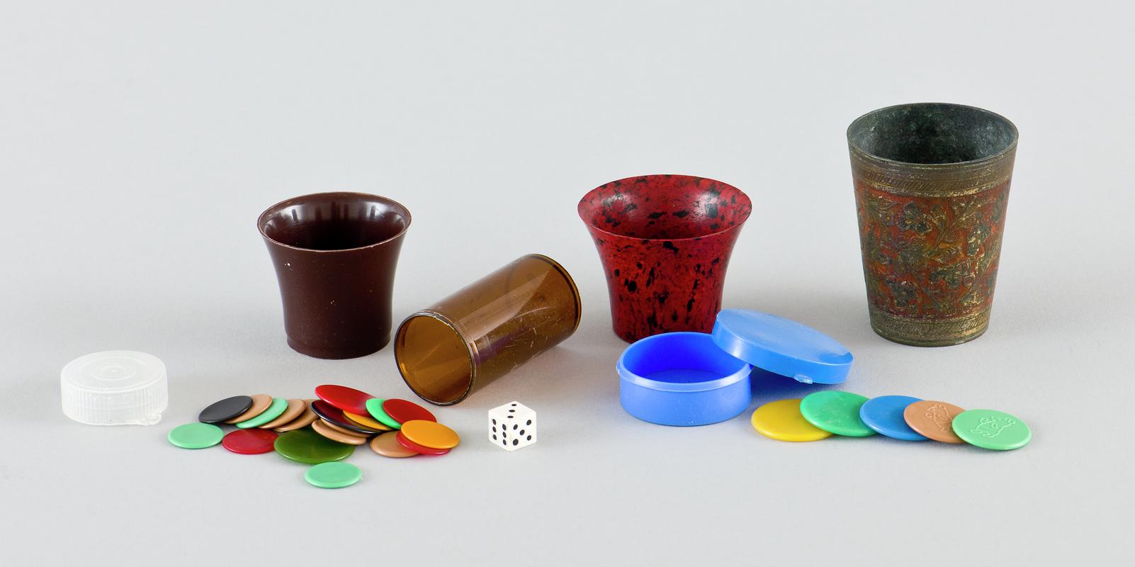 Tiddlywinks game and cups.  Twenty-one counters stored in brown plastic medication case (three larger than rest) along with a small plastic dice. Five counters (two a smaller design with bear motif) stored in blue plastic case with bear motif on interior base.   Large metal cup with raised floral design around body on red background. Medium sized red and black marble effect plastic cup. Smallest cup is plain brown plastic.