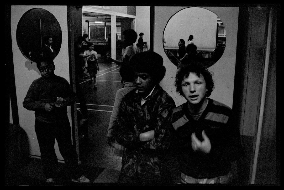 Group of teenagers, standing by an open door. There is another group dancing in the background, Butetown Youth Club.