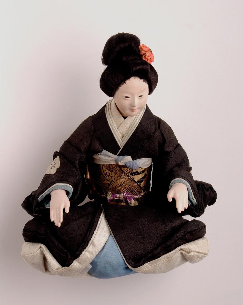 &#039;Sakura&#039; type Japanese doll, in kneeling position; head, forearms and lower legs of wood coated with gofum of a slightly fleshy colour; intricate coiffure, with artificial flower and tassel; rectangular holes for eyes; costume of black kimono; with usual