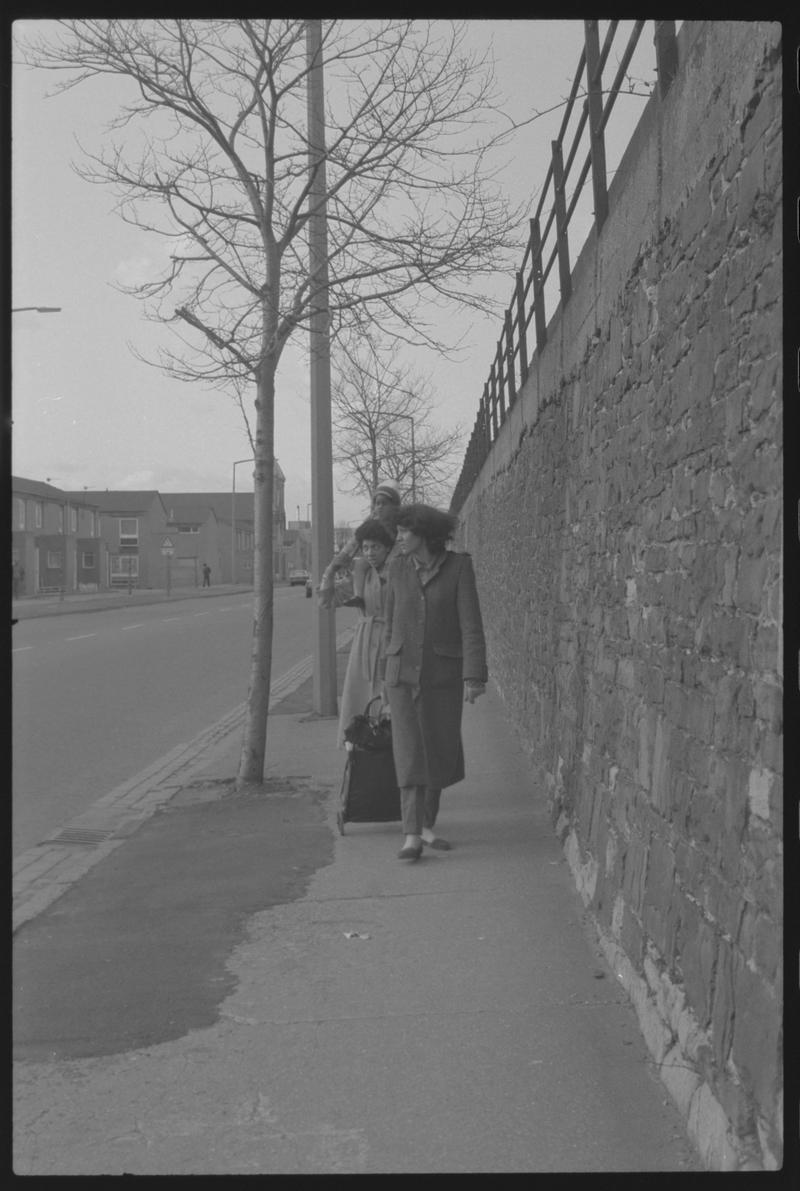 People walkiing along pavement by railway embankment wall, opposite shops on Bute Street.