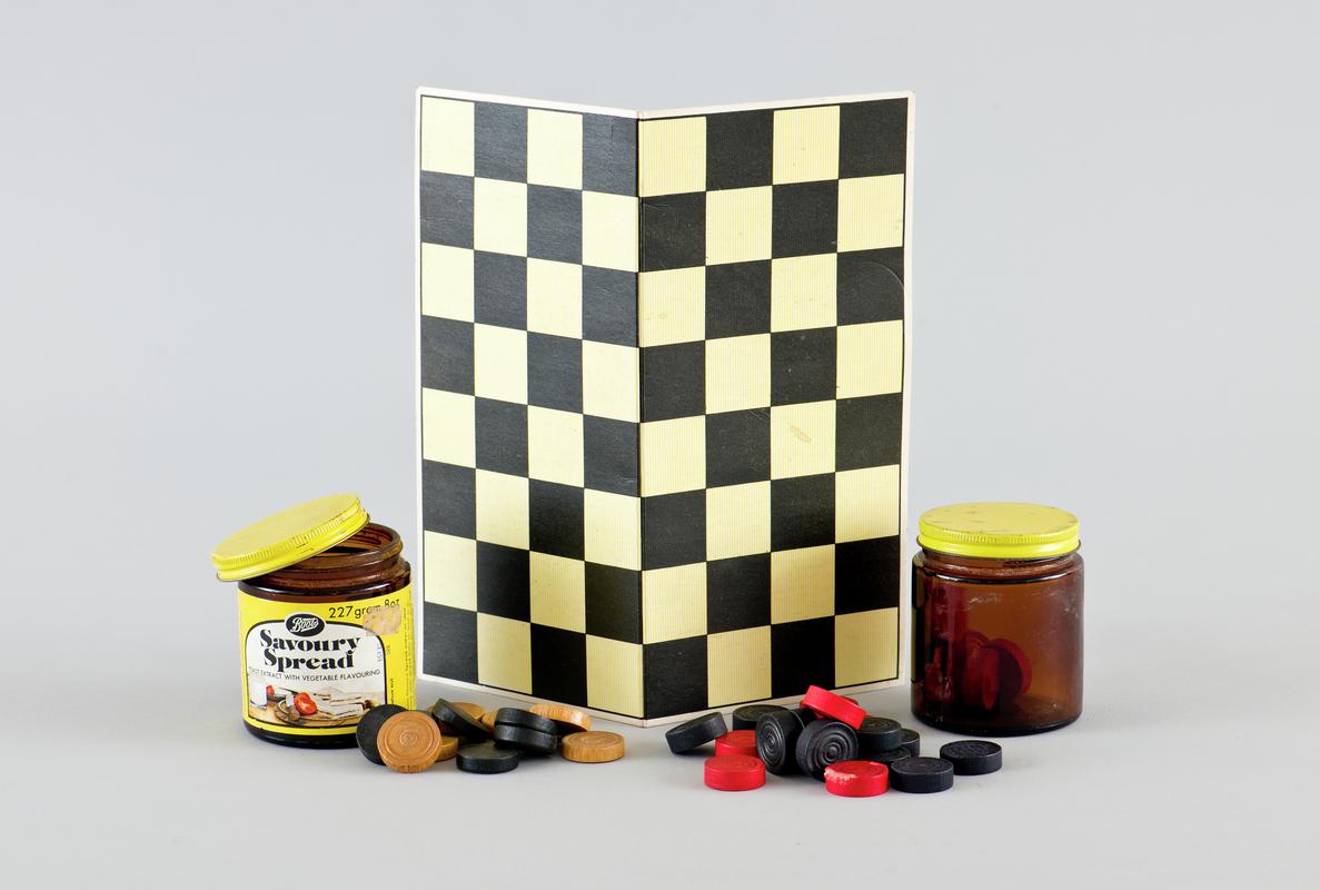 Cardboard draughts board, and two 8oz &#039;Boots Savoury Spread&#039; jars containing two sets of draught pieces: one set of black and tan draughts (11 of each) and one set of black and red draughts (12 of each and some counters damaged).