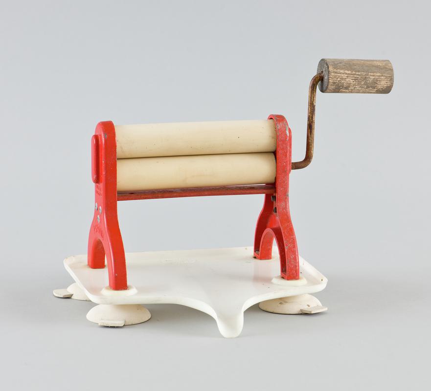 Tabletop clothes mangle called the &#039;Portair Limpet&#039;. Cream rubber (?) rollers within a red metal frame with well worn wooden handle to operate, sitting in a white plastic water tray with spout that can direct water into bowl. Tray sits on four cream rubber feet that can be stuck to flat surface. Product title embossed on side of two red metal uprights holding rollers.