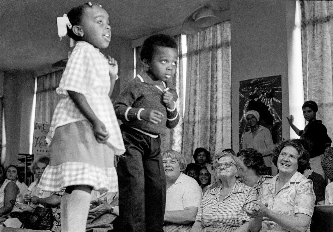 Young children on the catwalk pose for a fashion show in Butetown Community Centre, while the audience applaud. Cardiff, Wales