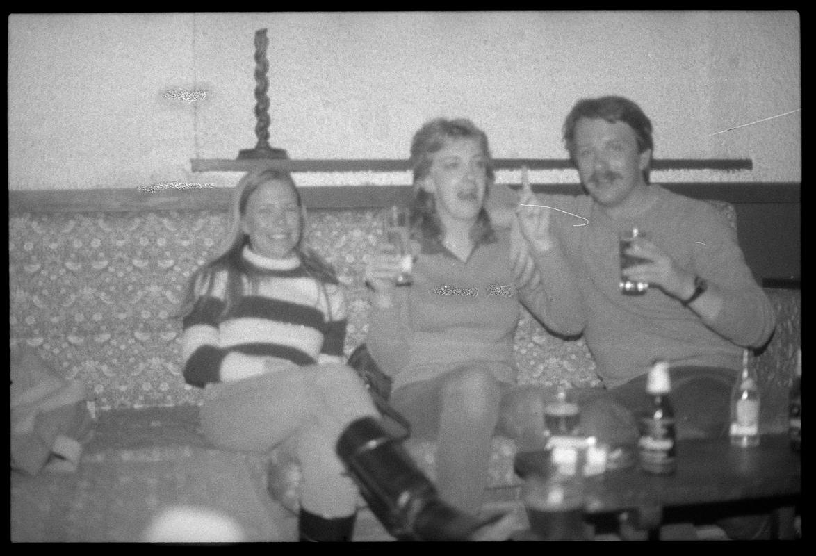 Three unidentified people sitting at table, outdoors. Possibly members of the TNT Group. Location unknown.