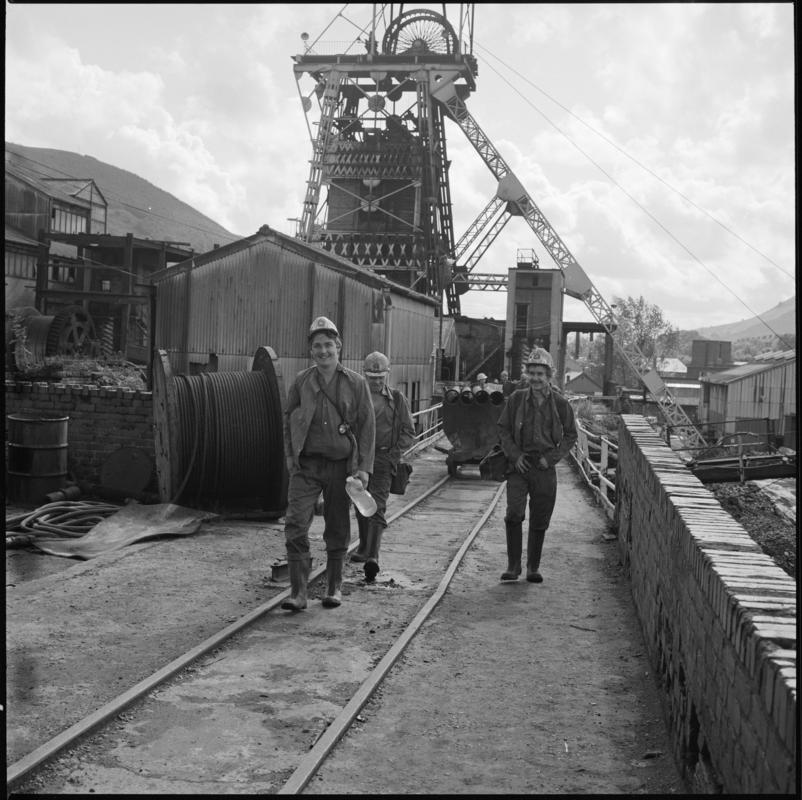 Black and white film negative showing a surface view of Merthyr Vale Colliery with three men in the foreground, 1976.  &#039;Merthyr Vale 1976&#039; is transcribed from original negative bag.
