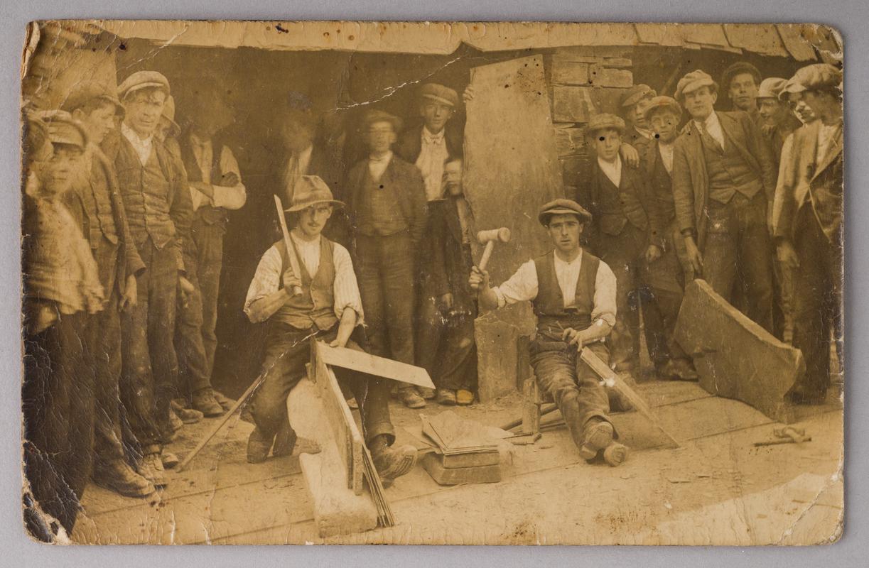 Aged photographic print showing quarrymen outside one of the gwaliau (an open-fronted shed where slates were split and trimmed) at a slate quarry (believed to be Penrhyn Quarry).
