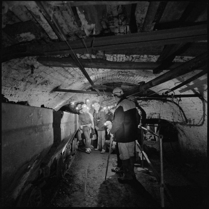 Black and white film negative showing visitors to Big Pit Mining Museum with guide at a junction near pit bottom, 1983.
