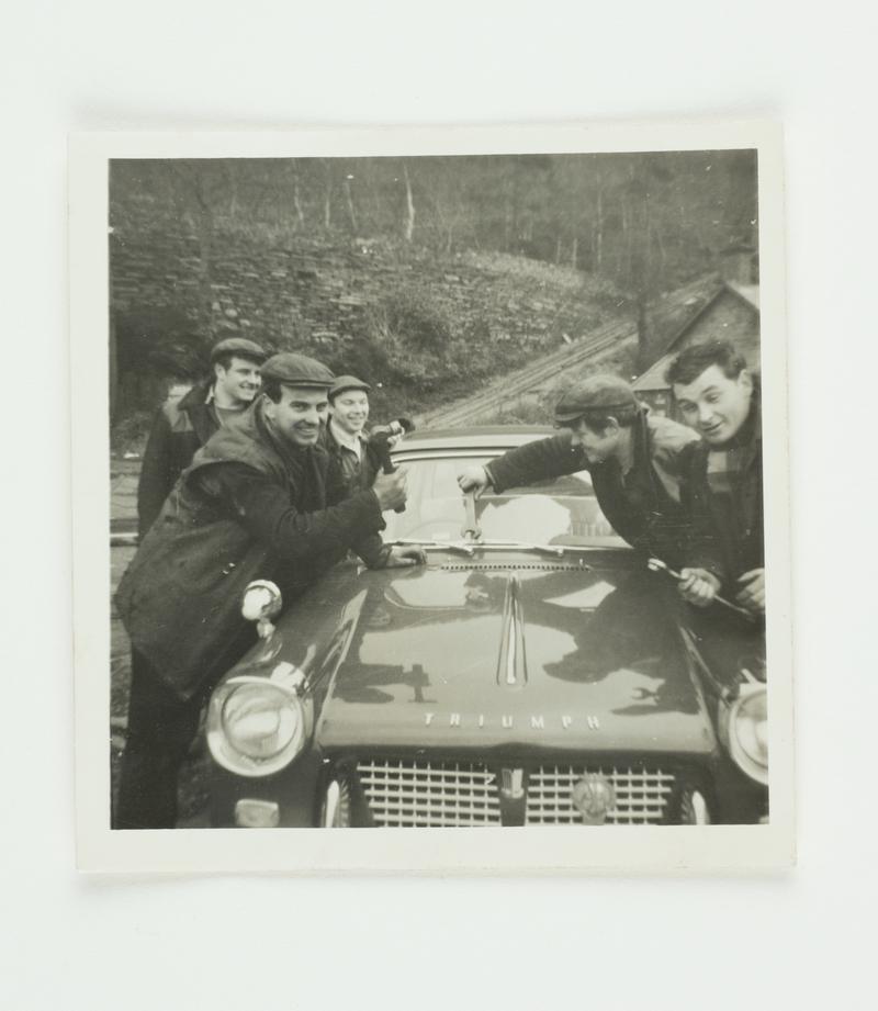 Five members of staff of the Dinorwig Quarry Workshop situated around a Triumph Herald motor car. On the left is Alun Roberts (fitter), Arfon Jones (fitter) and Vernon Griffiths (air fitter). On the right is Billy Williams (fitter) and Ken Jones (fitter). Taken at Gilfach Ddu, Llanberis.