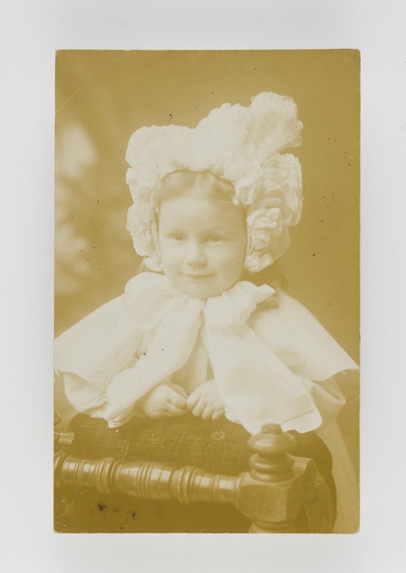 Head &amp; shoulders photograph of a young girl in bonnet looking over chair. (1900s)