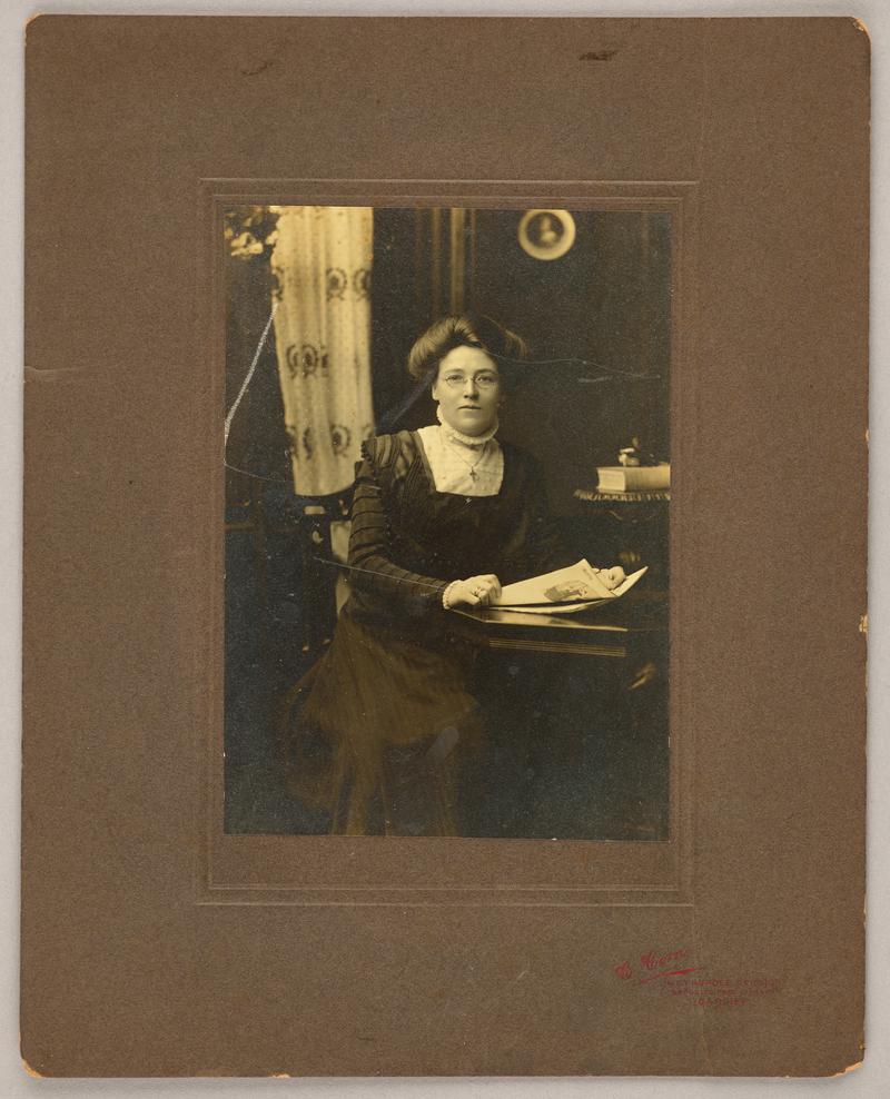 Portrait of unknown woman sitting at a table reading. Mounted on brown card with photographers details printed bottom right.