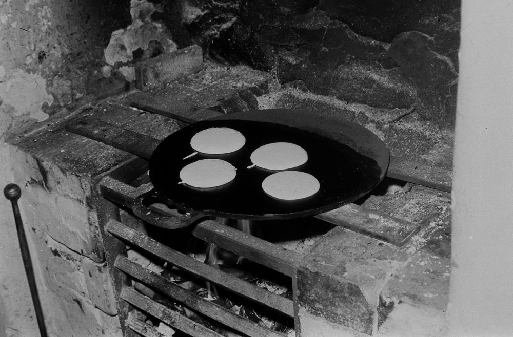 Black-and-white photograph of four pancakes cooking on a griddle over an old-fashioned brick fireplace.