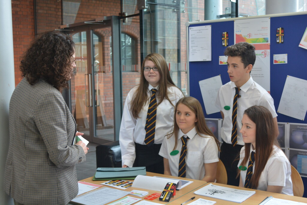 A women talking to a group of older school children who are sat behind a table.