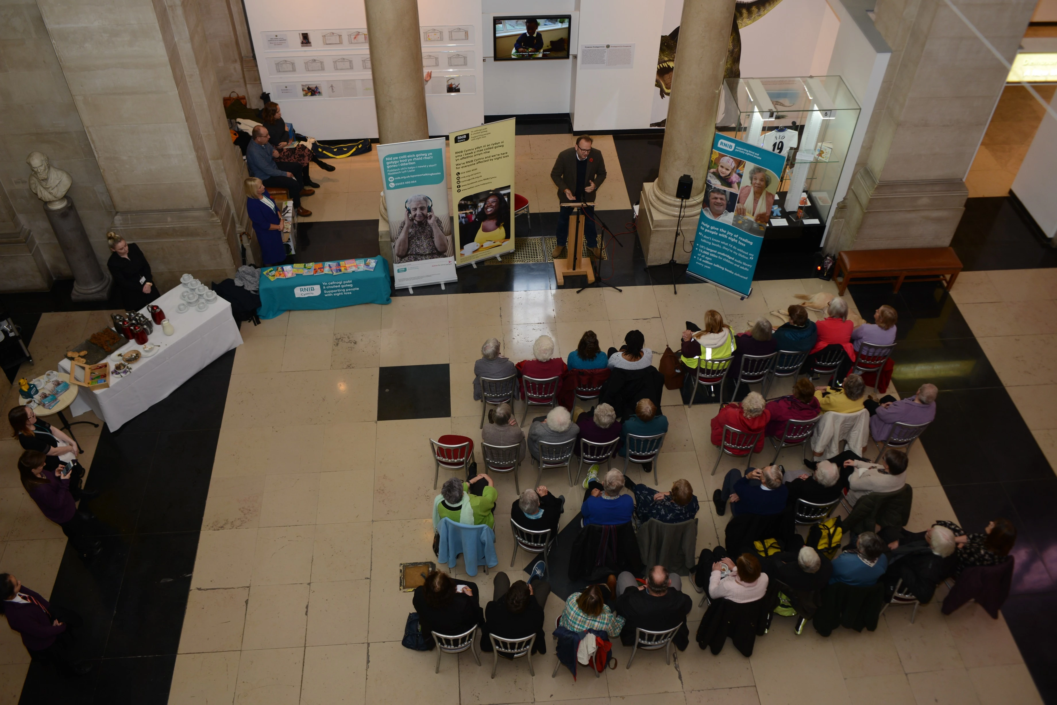 Overhead photograph of a crowd listening to a man speak in the main hall of the musuem