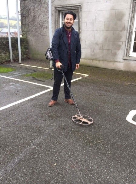 A man is holding a metal detector in a court yard with a smile on his face