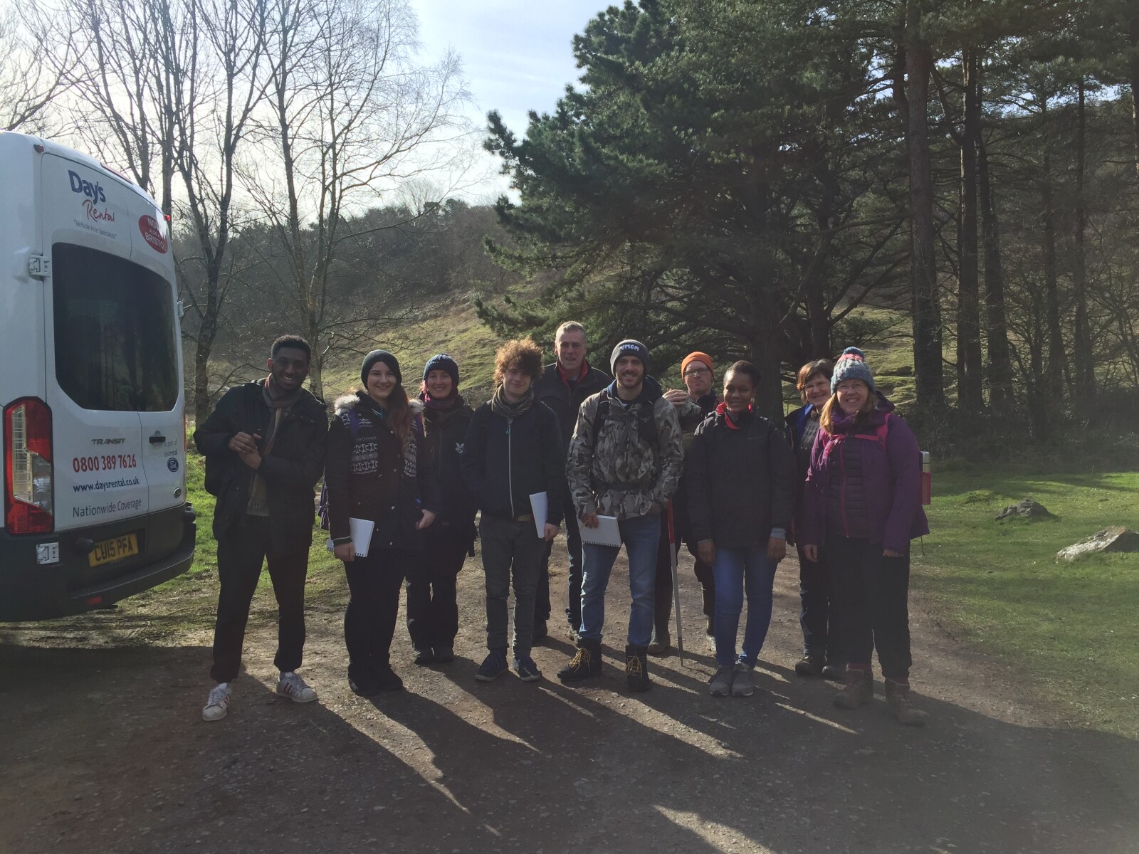 The images shows a variety of people at the bottom of a hill in a woodland area; they are about to go on a long walk. 