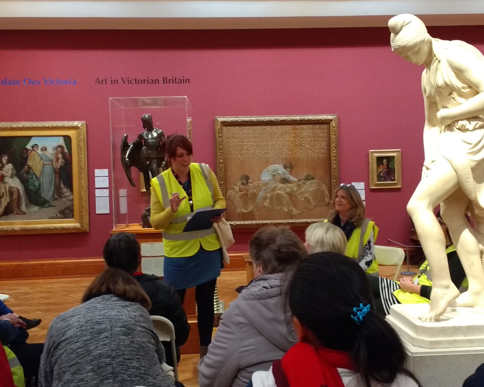 A crowd of people sitting in the Victorian Art gallery at National Museum Cardiff, listening to a tour guide