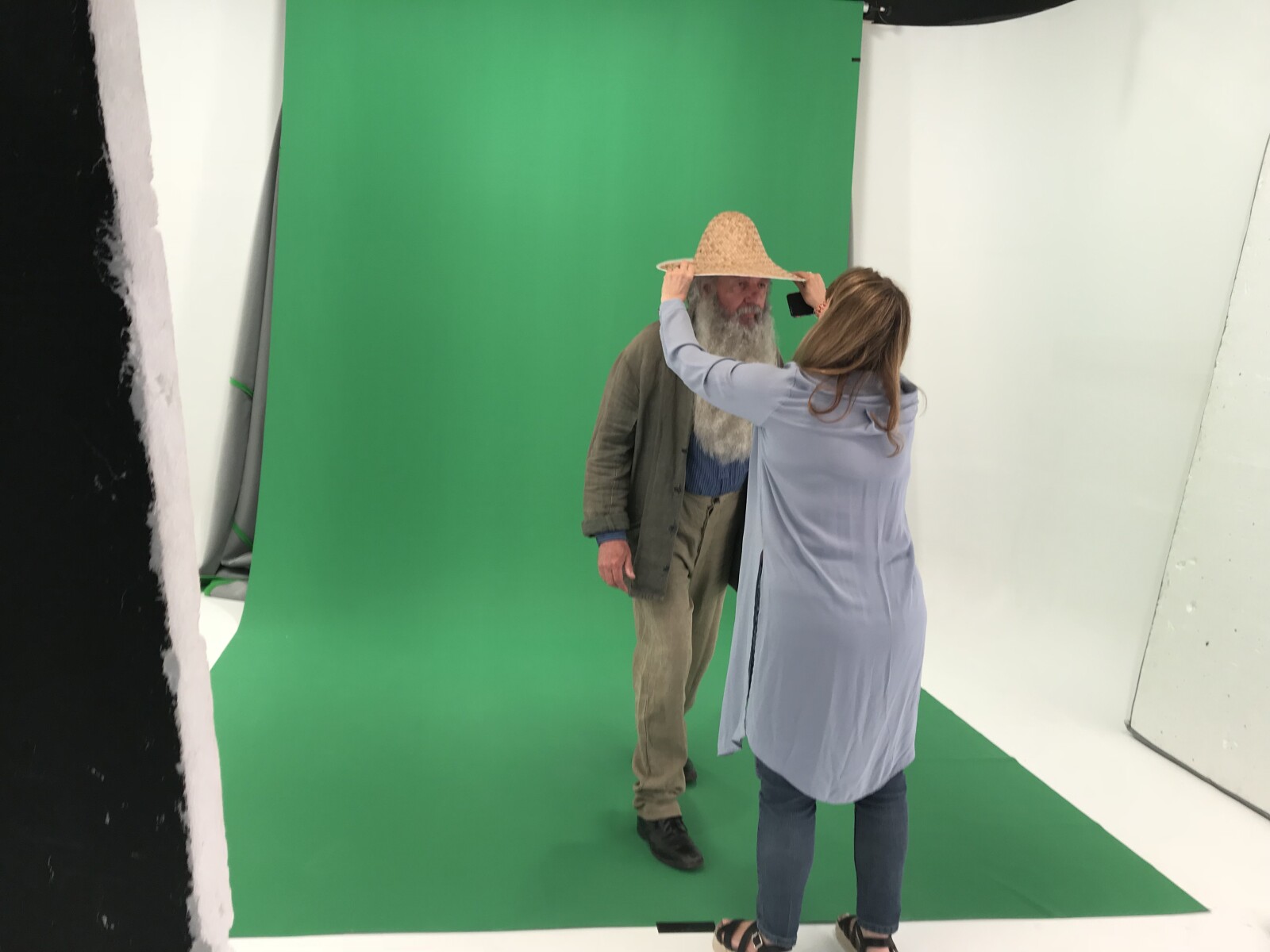 Actor dressed as the artist Monet standing in front of a green screen having his hat adjusted by a make up artist 