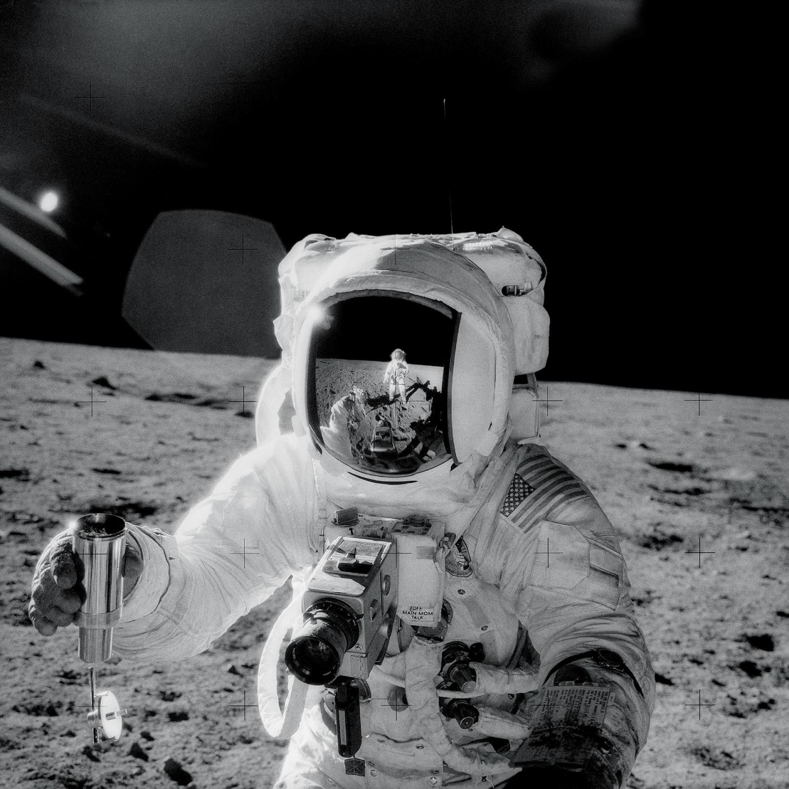 One of the astronauts on the Moon’s surface is holding a container of lunar soil. The other astronaut is seen reflected in his helmet.