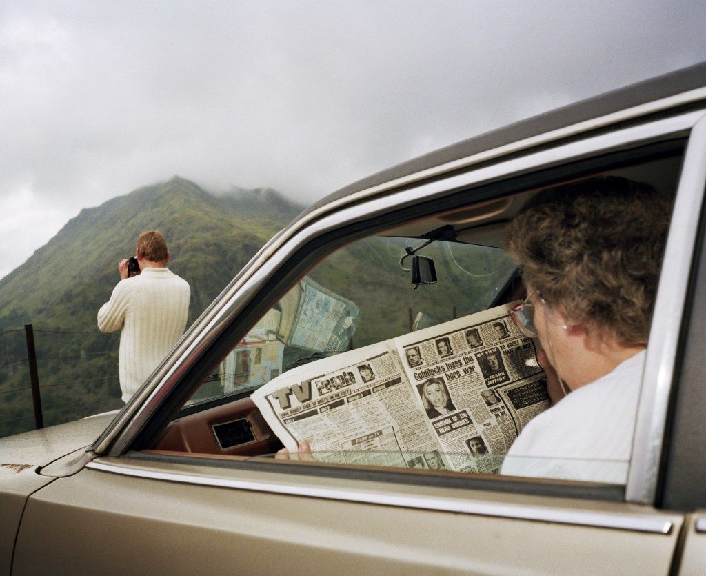 A photograph of a man looking out towards the mountains while a woman reads the paper in a car