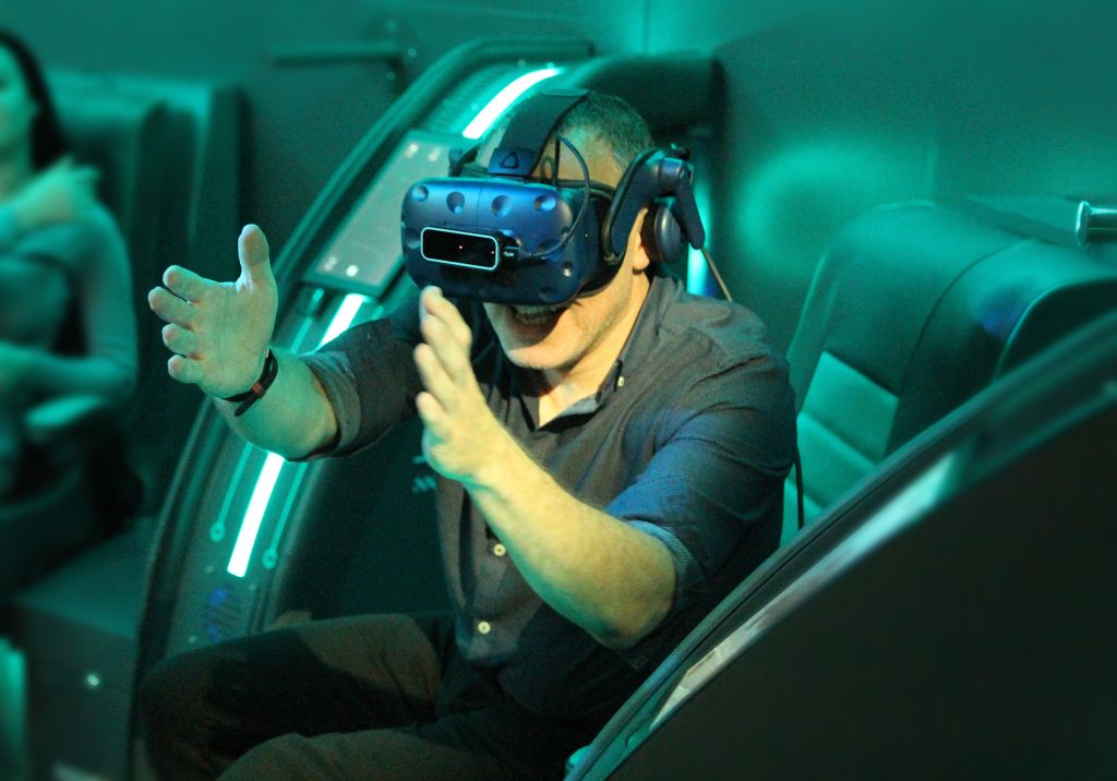 a young man in a rumble seat wearing a VR headset and with his arms outstretched 