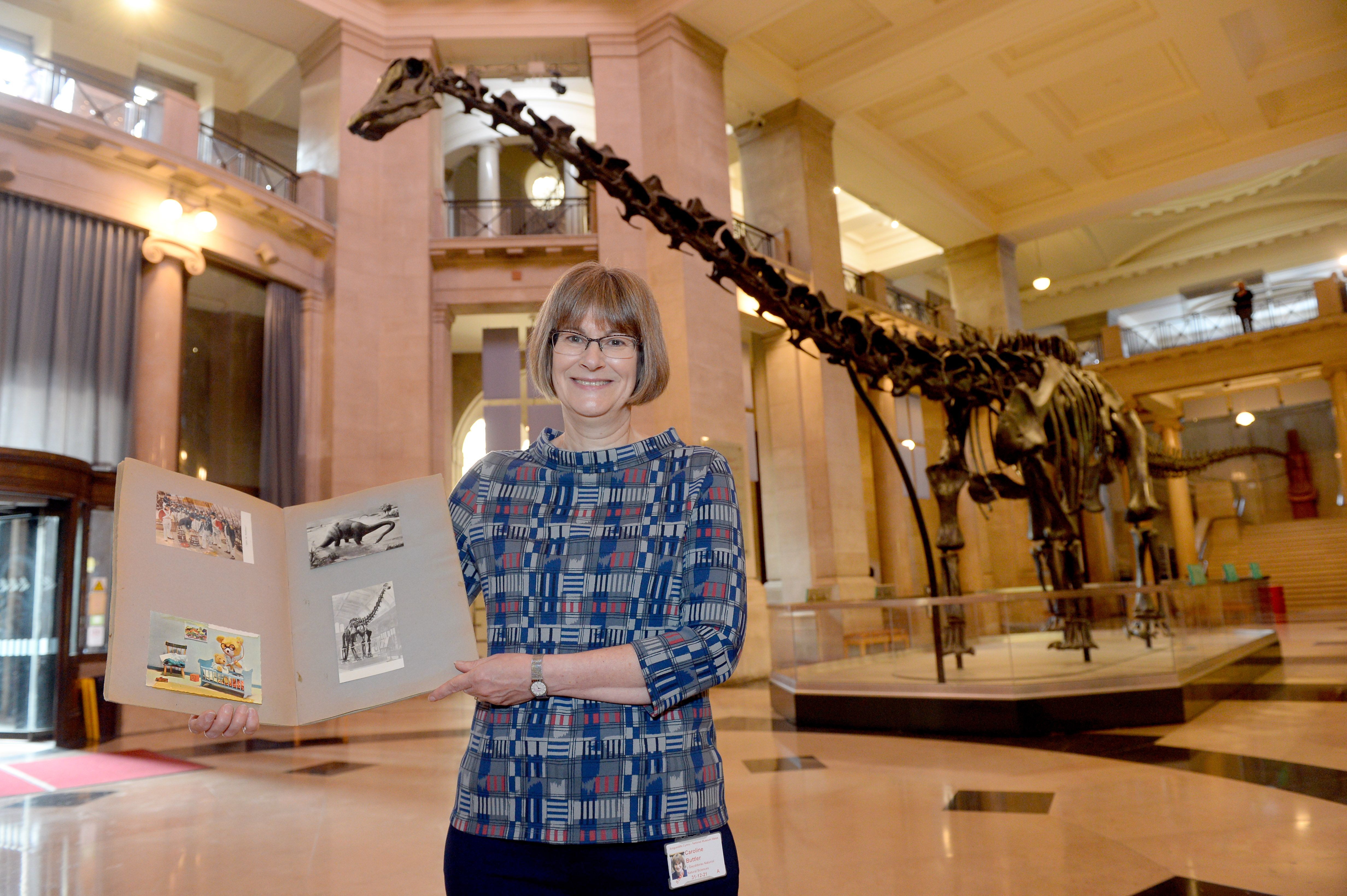 Dr Caroline Buttler with her scrapbook in National Museum Cardiff's main hall with Dippy in the background
