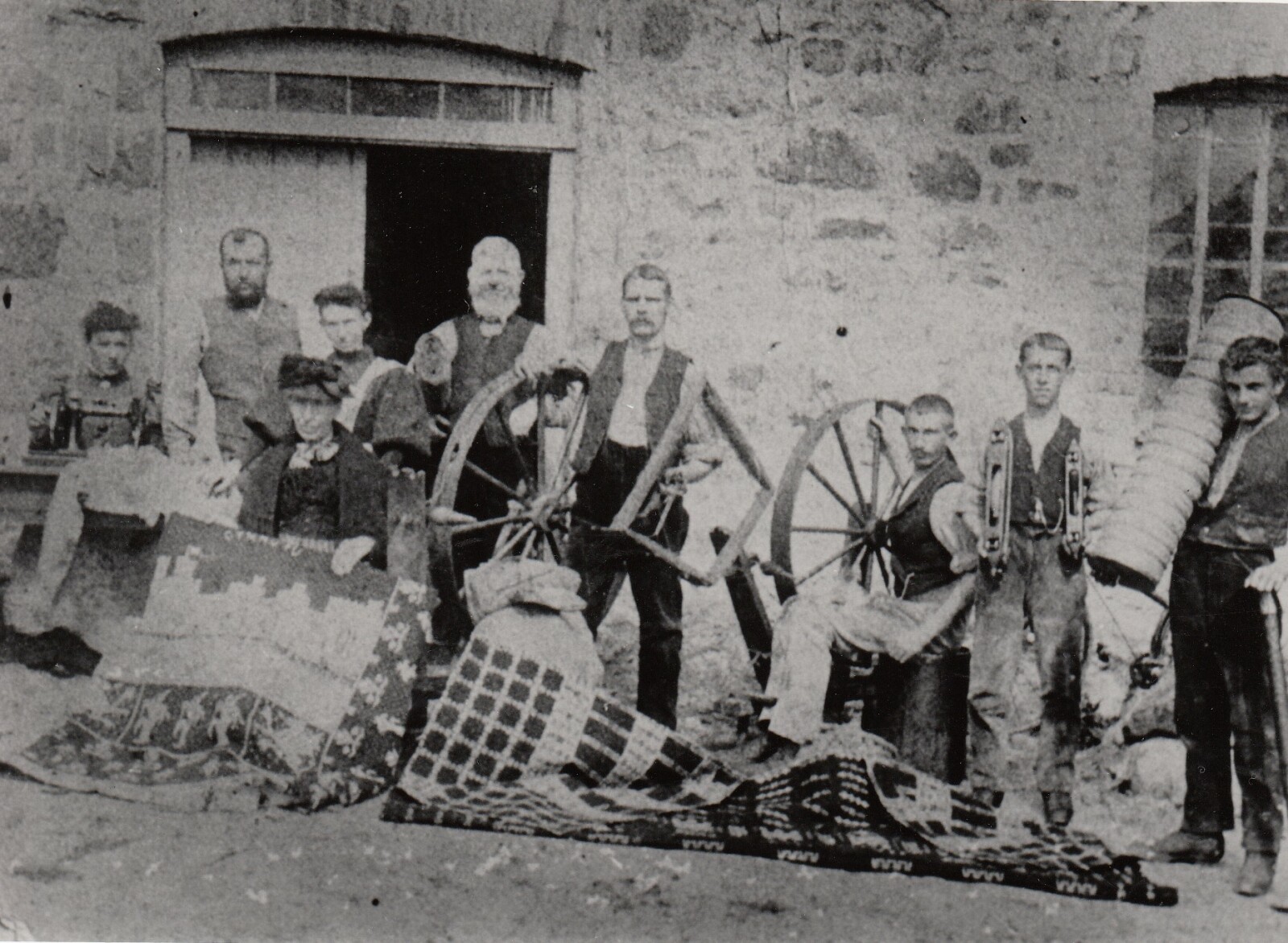Black and white photograph showing nine workers outside Pwllheli Woollen Mill with blankets, spinning wheels and tools of their trade