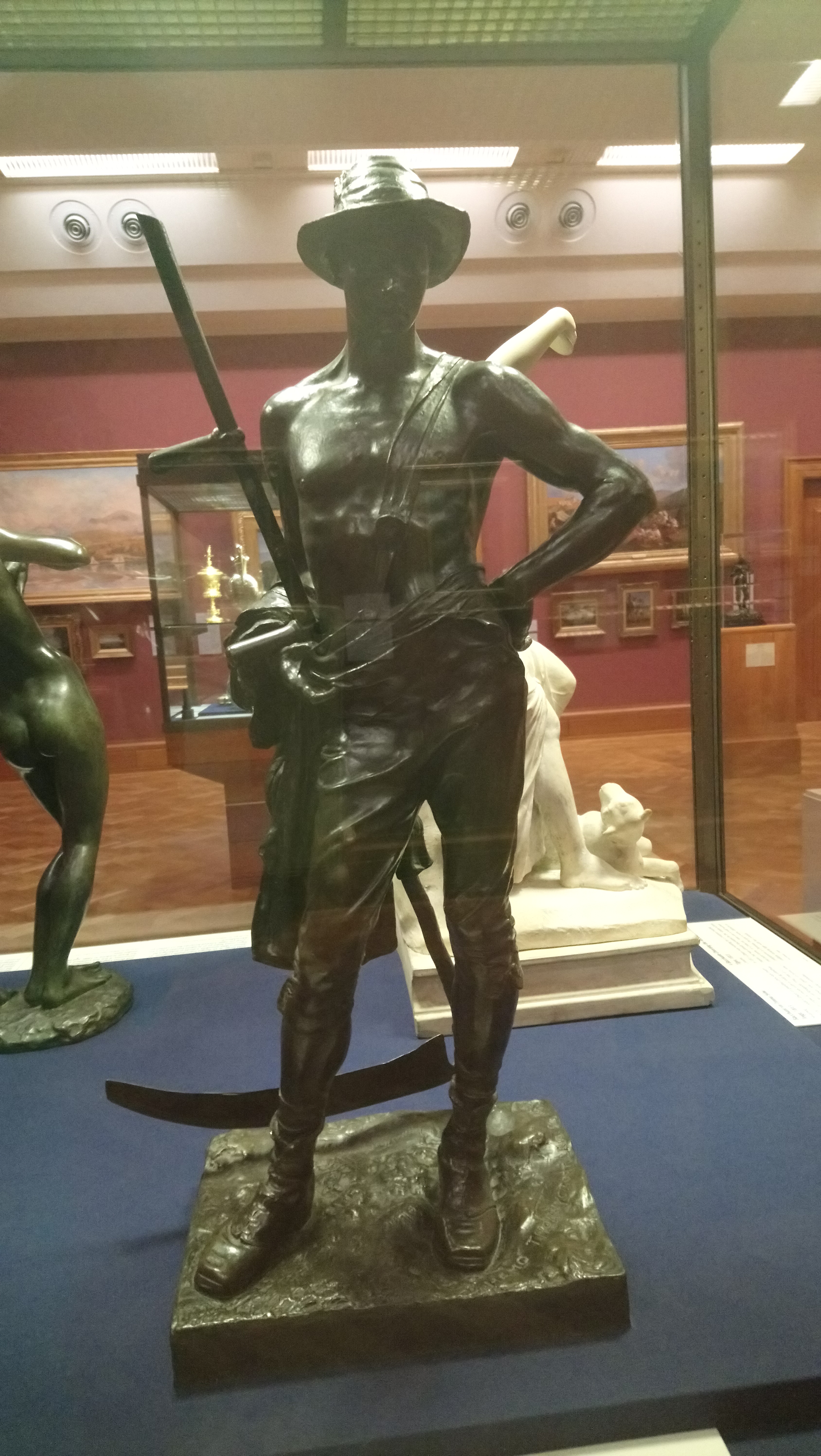 A bronze sculpture of a semi-nude male figure wearing a hat and holding a scythe