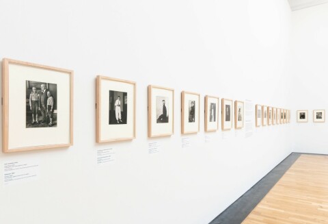 A photograph of the ARTIST ROOMS: August Sander exhibition, showing a series of square black and white portrait photographs in a line on a blank white wall