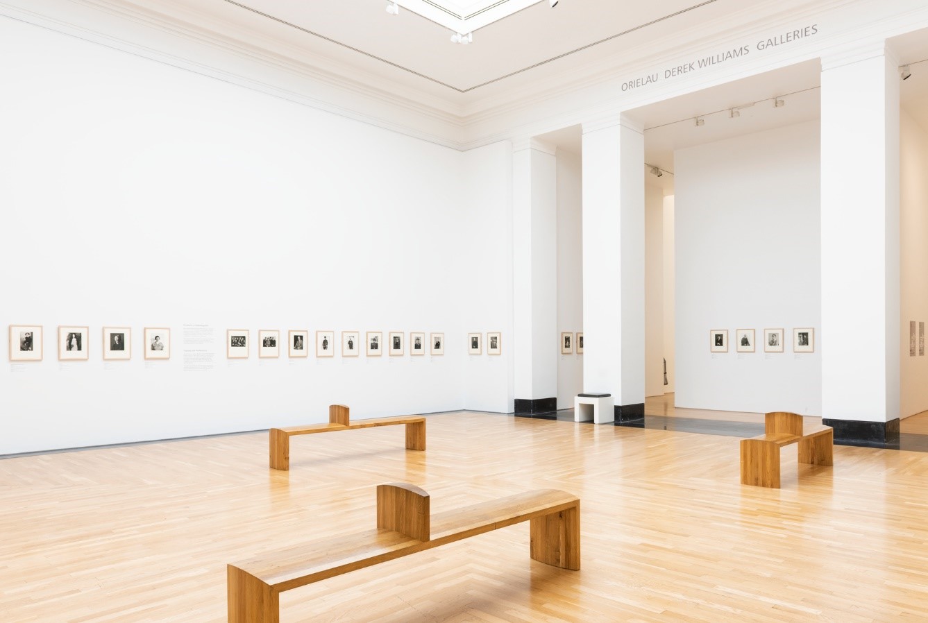A photograph of the ARTIST ROOMS: August Sander exhibition, showing the entire gallery with small photos on the walls and wooden benches in the middle