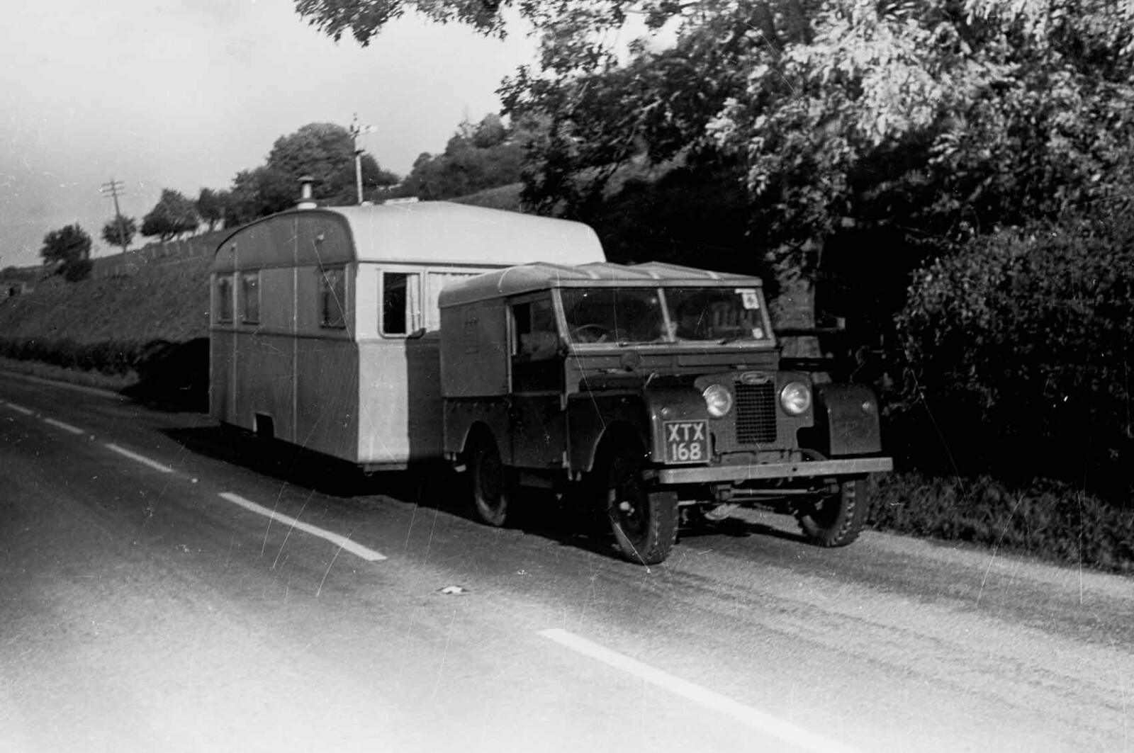 Black & white photograph of a Land Rover towing a caravan on a country road. 