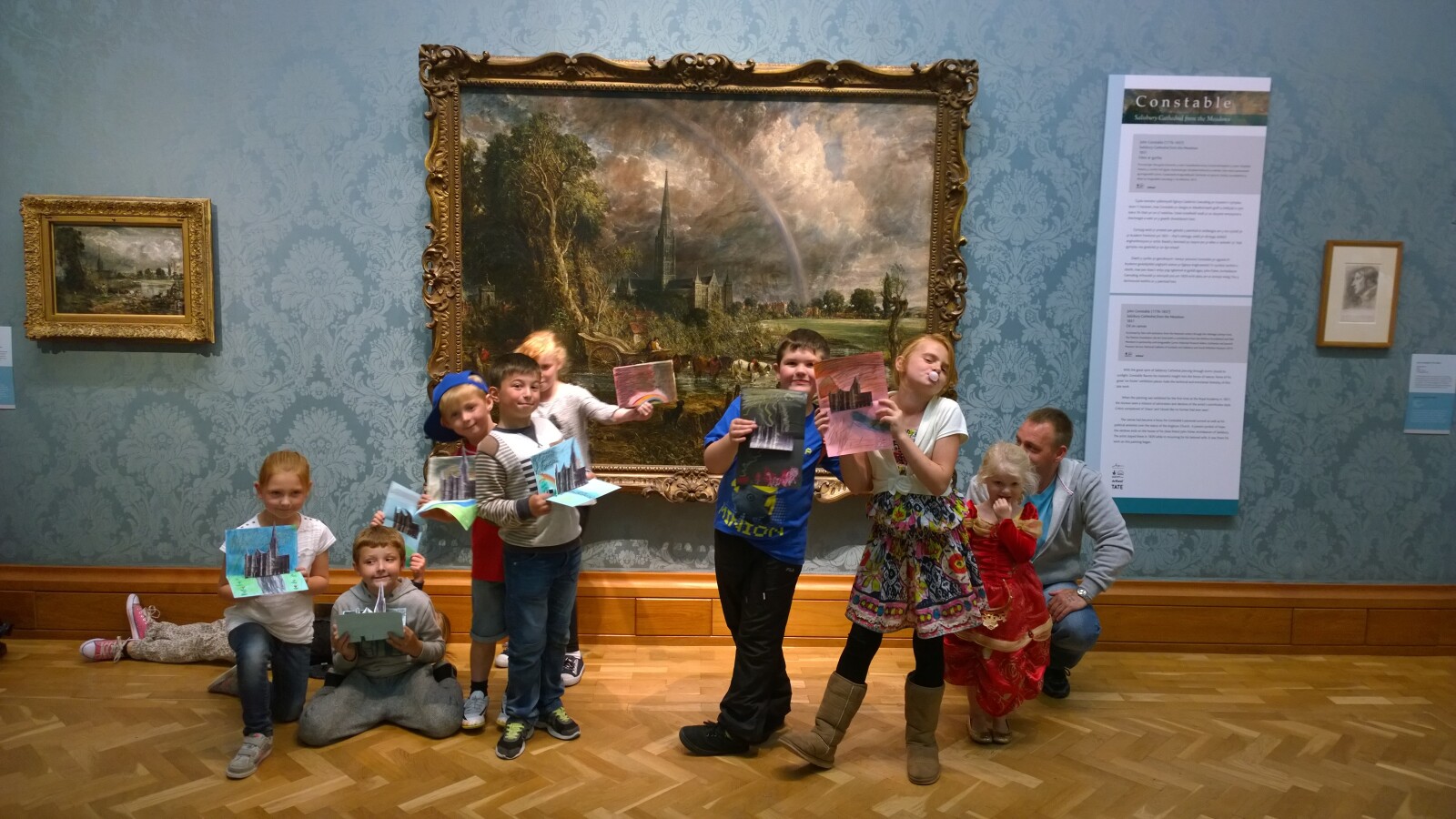 A group of young children standing in front of a painting, holding up their artworks